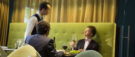 Un restaurante con un asiento tapizado de color verde, un camarero y una mesa de comedor con copas de vino y platos.
