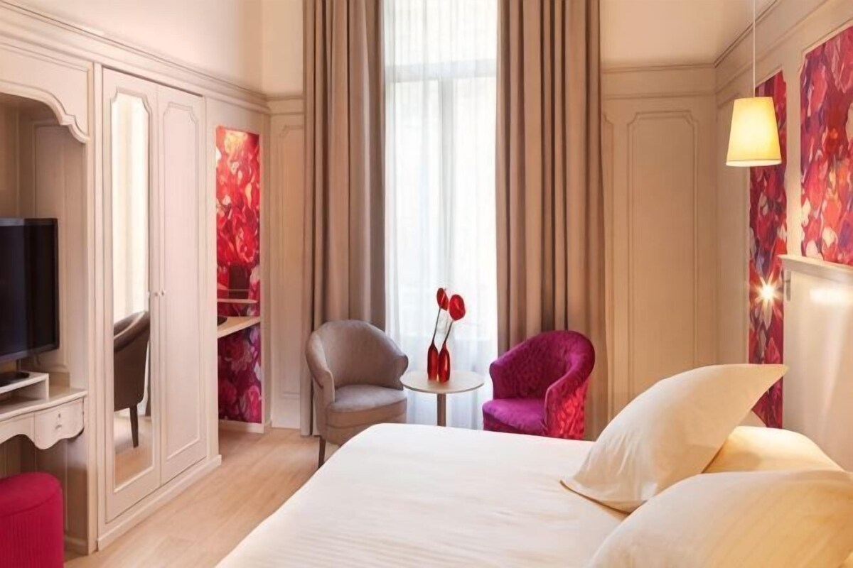 A hotel room with a bed, a television, a desk, a chair, a small table, and a red flower vase.