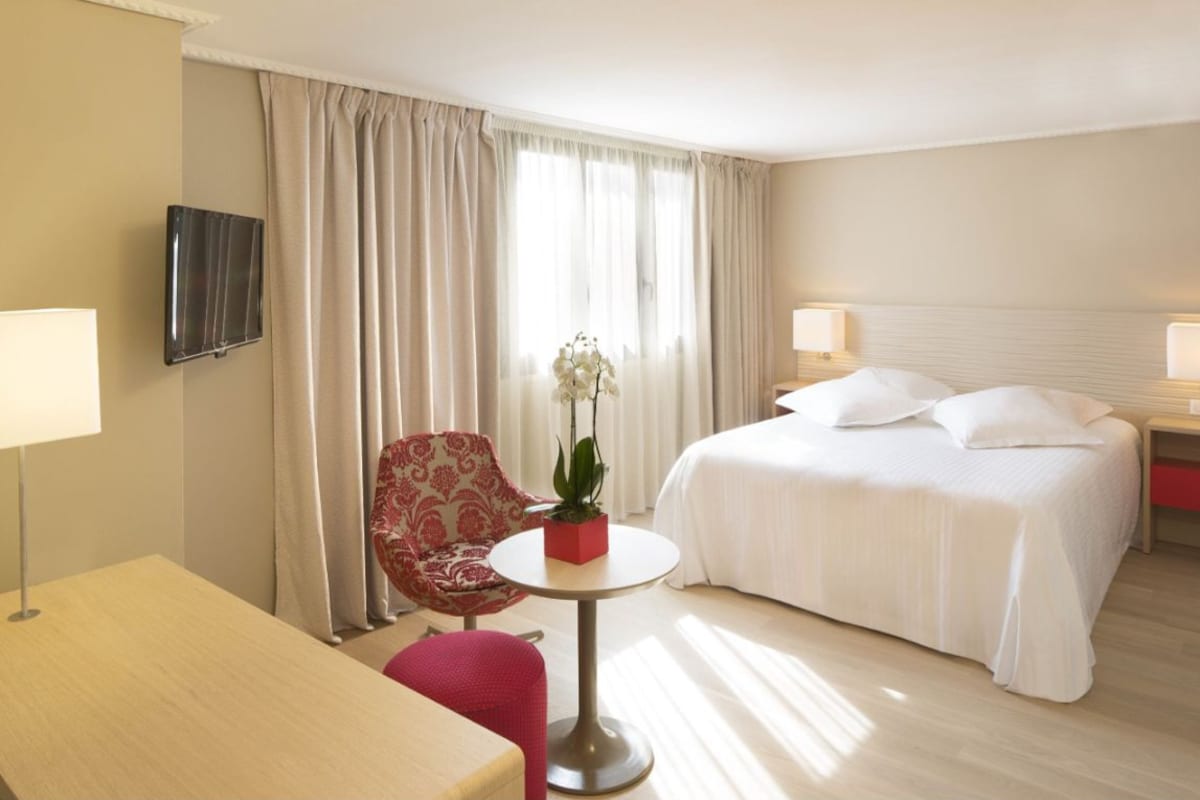 A hotel room with a bed, a chair, a small table with a plant, a TV mounted on the wall, and a lamp on the bedside table.