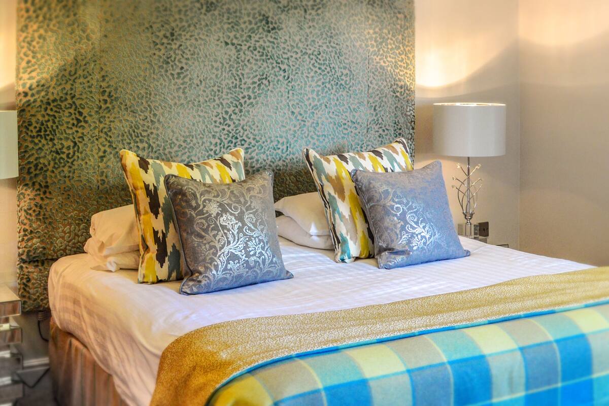 Una cama bien hecha con almohadones estampados y una colcha a cuadros, una lámpara de noche y una pared con textura.