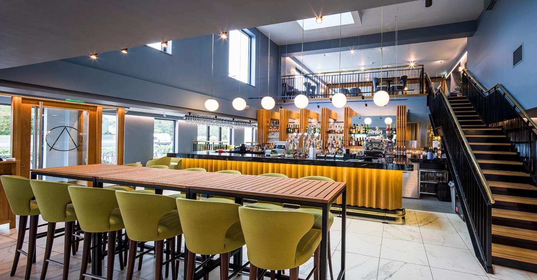 A modern bar area with a wooden bar counter, yellow bar stools, and a staircase leading to an upper level.
