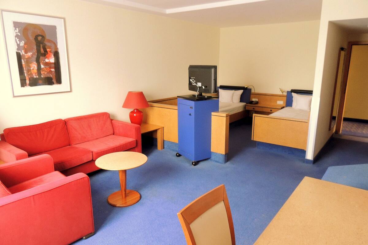 A compact hotel room with a red sofa, a small round table, a desk with a computer, a bed, and a blue cabinet.