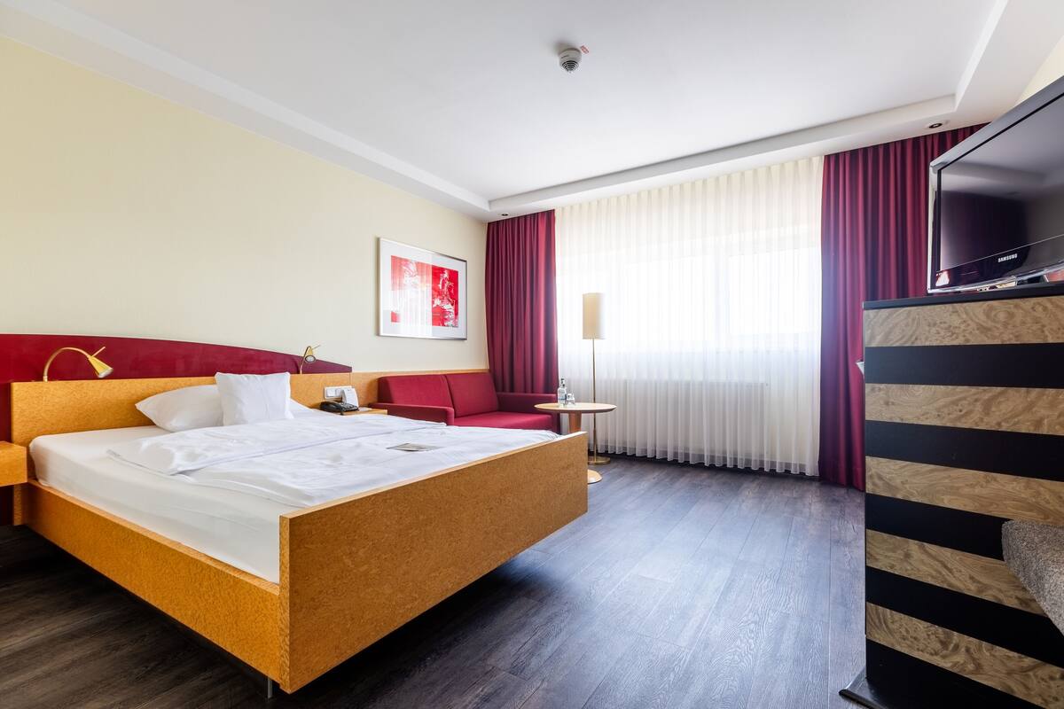 A hotel room with a large bed, a red sofa, a small round table, and a TV on a wooden stand.