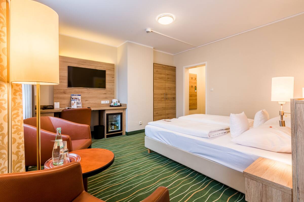 A hotel room with a bed, a desk with a flat-screen TV, a seating area with chairs, and a minibar.