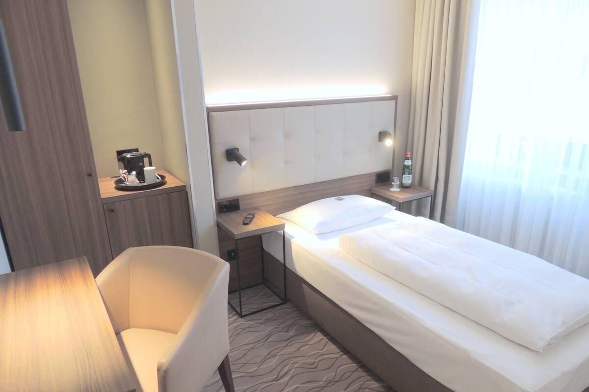 A hotel room with a bed, a desk, a chair, a nightstand, a remote control, and a bottle of water.