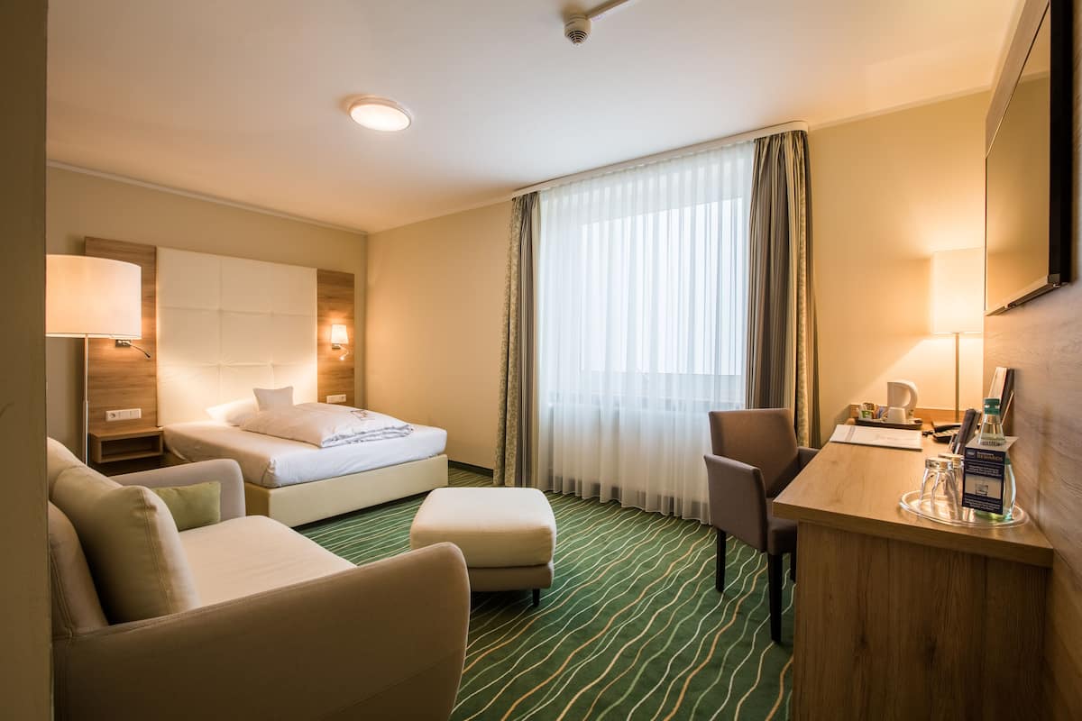 A hotel room with a bed, two armchairs, a desk, and a large window with curtains.