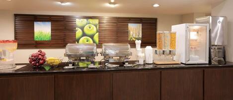 Zona de desayuno de hotel con refrigerador, máquinas de café y frutero.