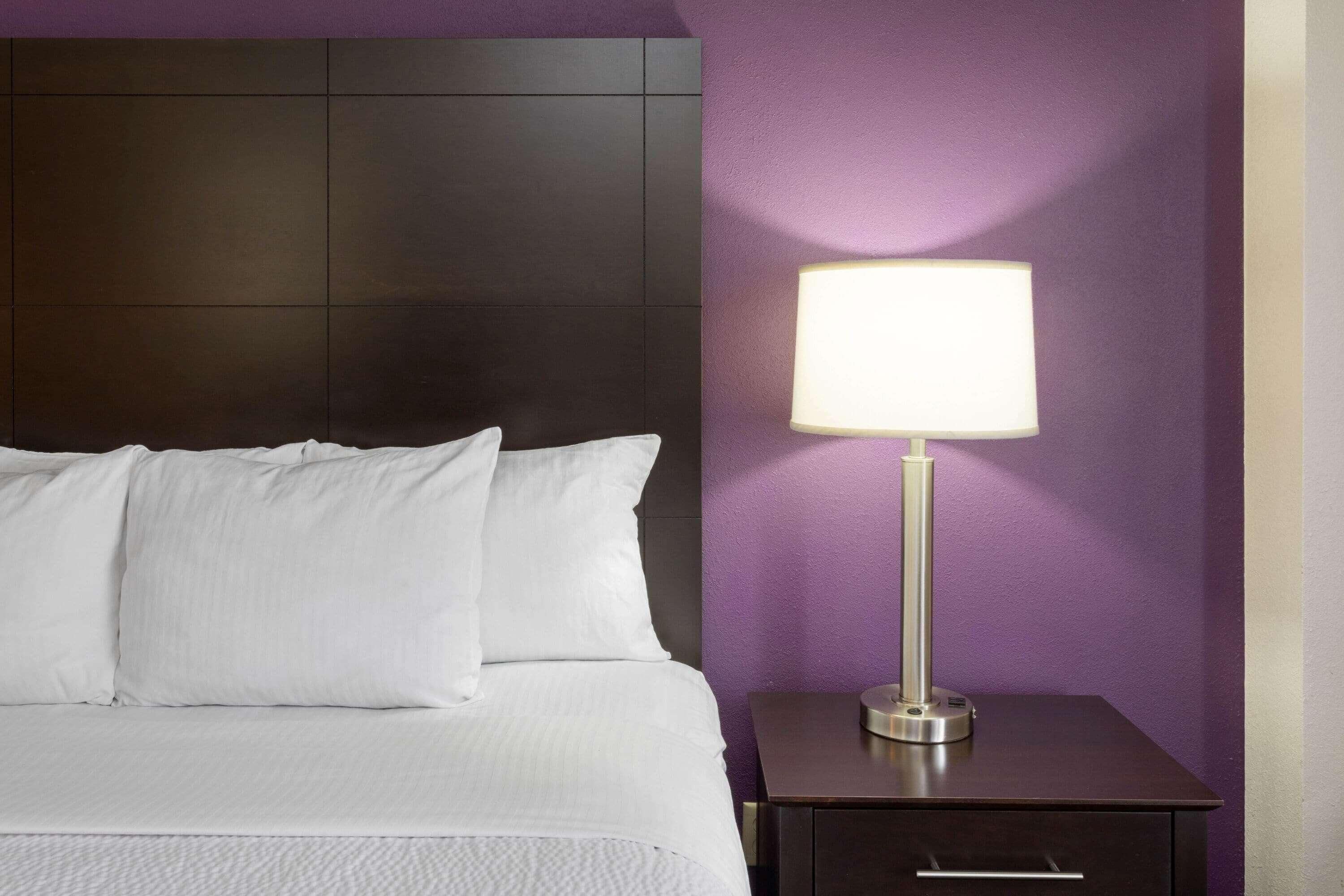 A neatly made bed with white linens, a dark headboard, a bedside table with a lamp, and a purple wall.