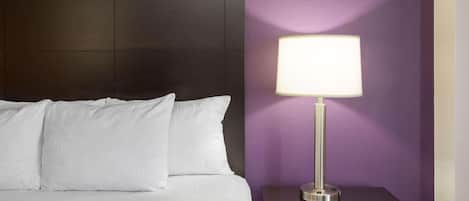 Una cama bien hecha con sábanas blancas, un cabecero oscuro, una mesita de noche con una lámpara y una pared morada.