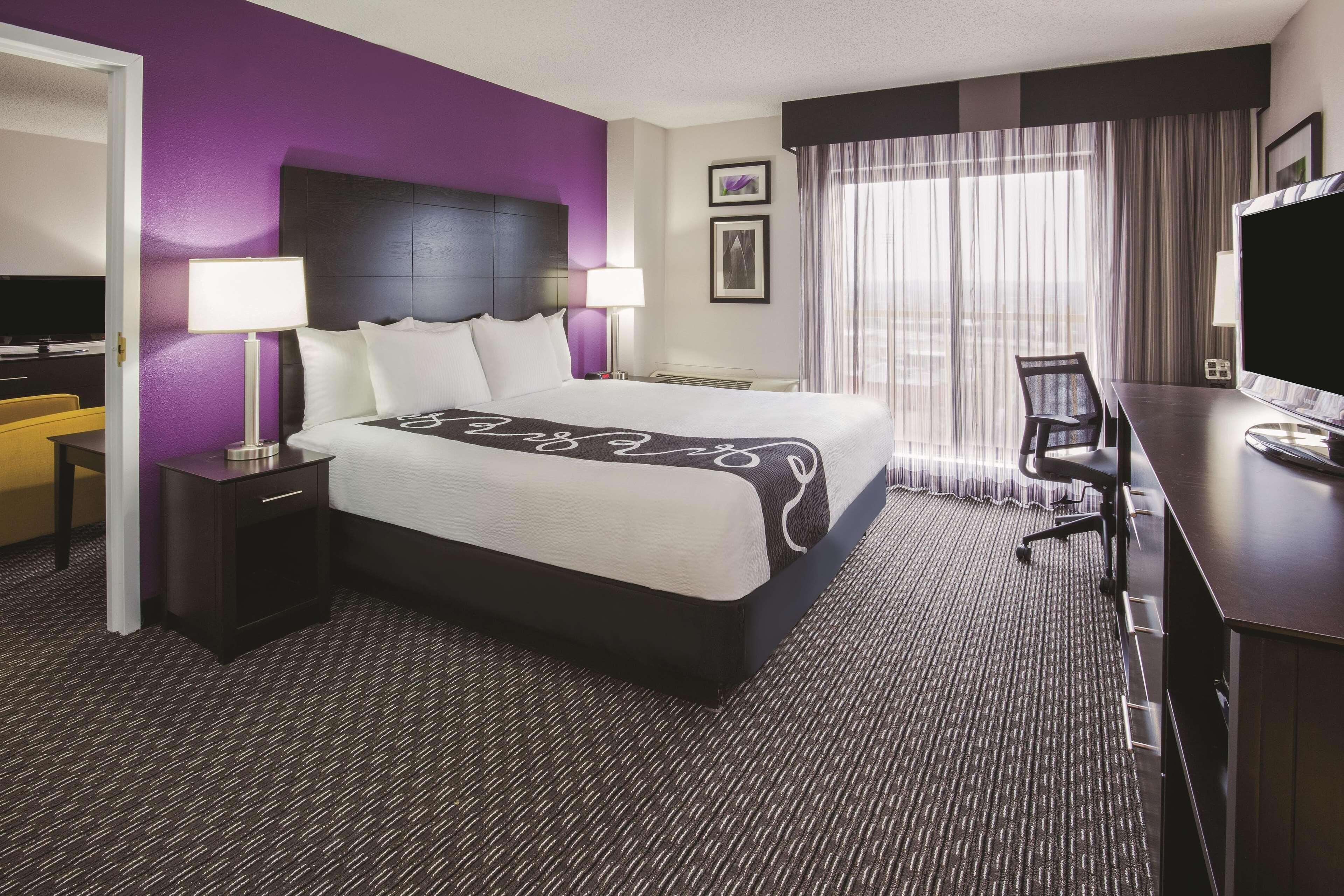 Habitación de hotel con una cama grande, dos mesitas de noche con lámparas, un escritorio con silla, un televisor y una ventana con cortinas.