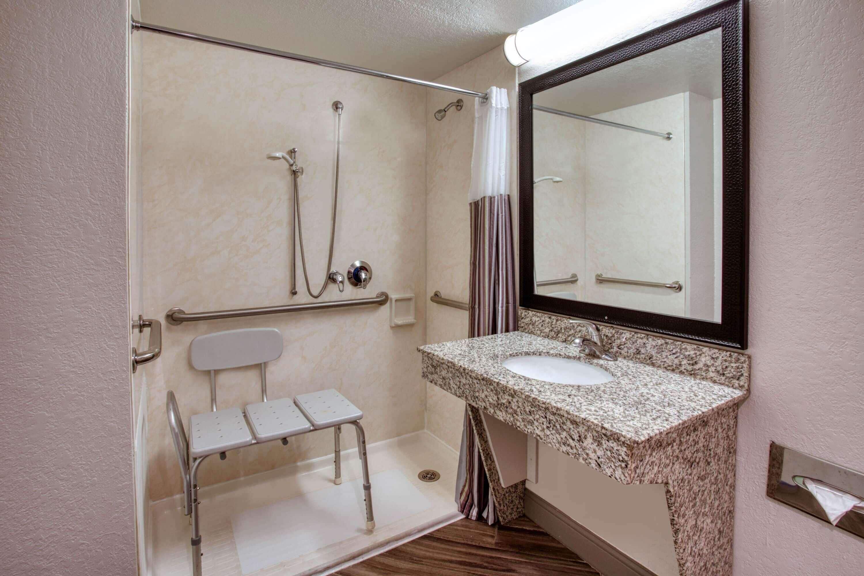 A bathroom with a shower, a sink, a mirror, and a wheelchair-accessible bench.