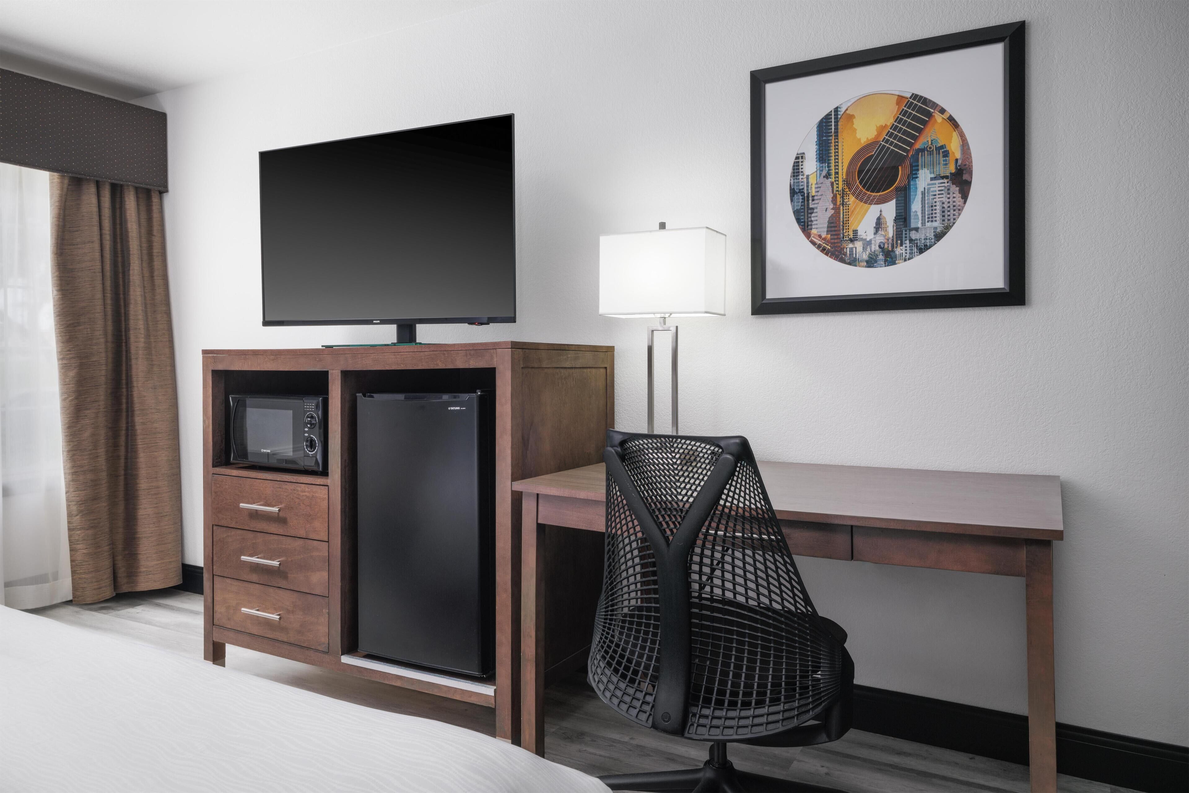 Habitación de hotel con cama, escritorio con lámpara, televisor, microondas y refrigerador.