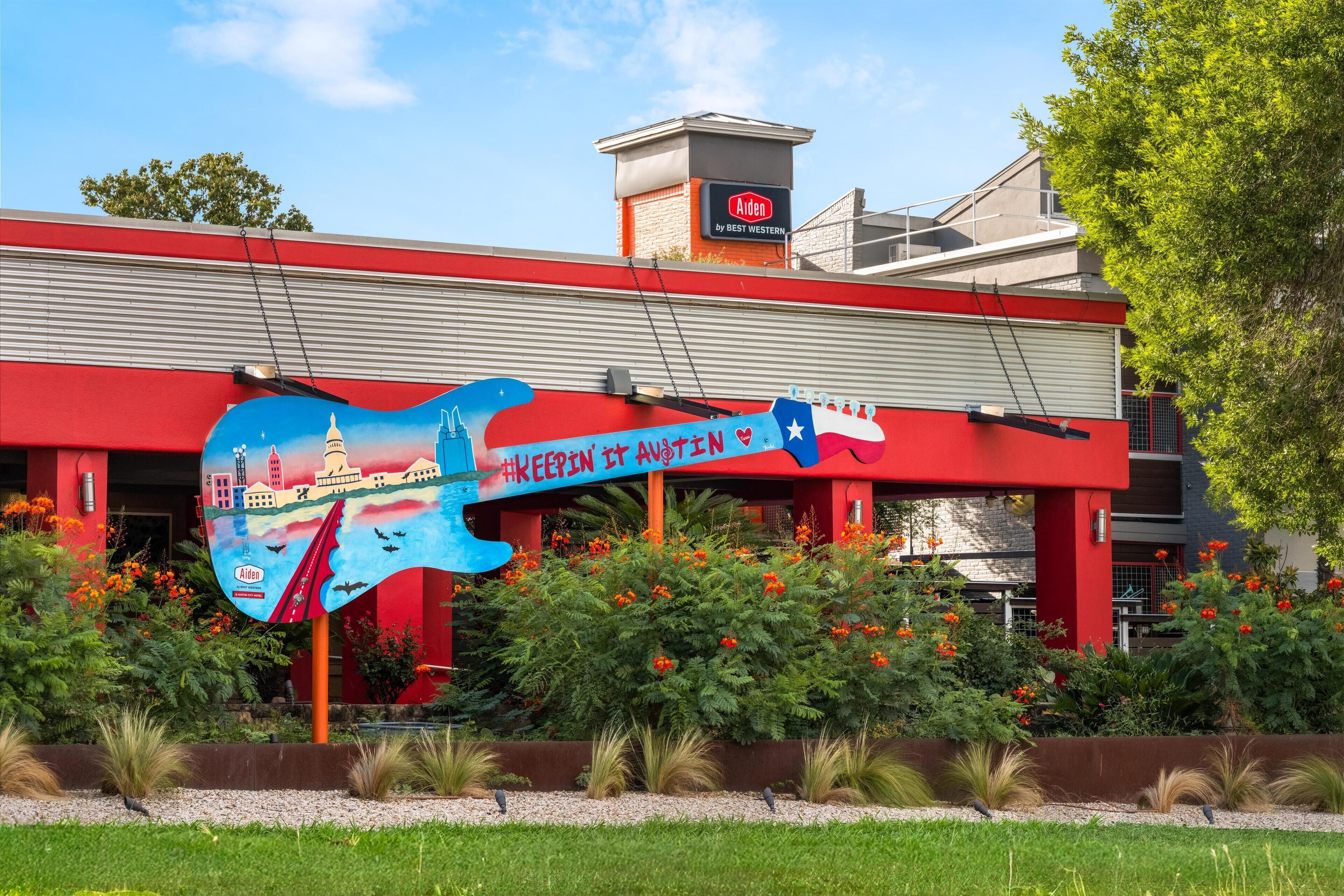 A building with a red facade, a large guitar-shaped sign with the text "KEEPIN IT ANOTIN" and "Aiden by Best Western" on the roof, surrounded by greenery and flowers.