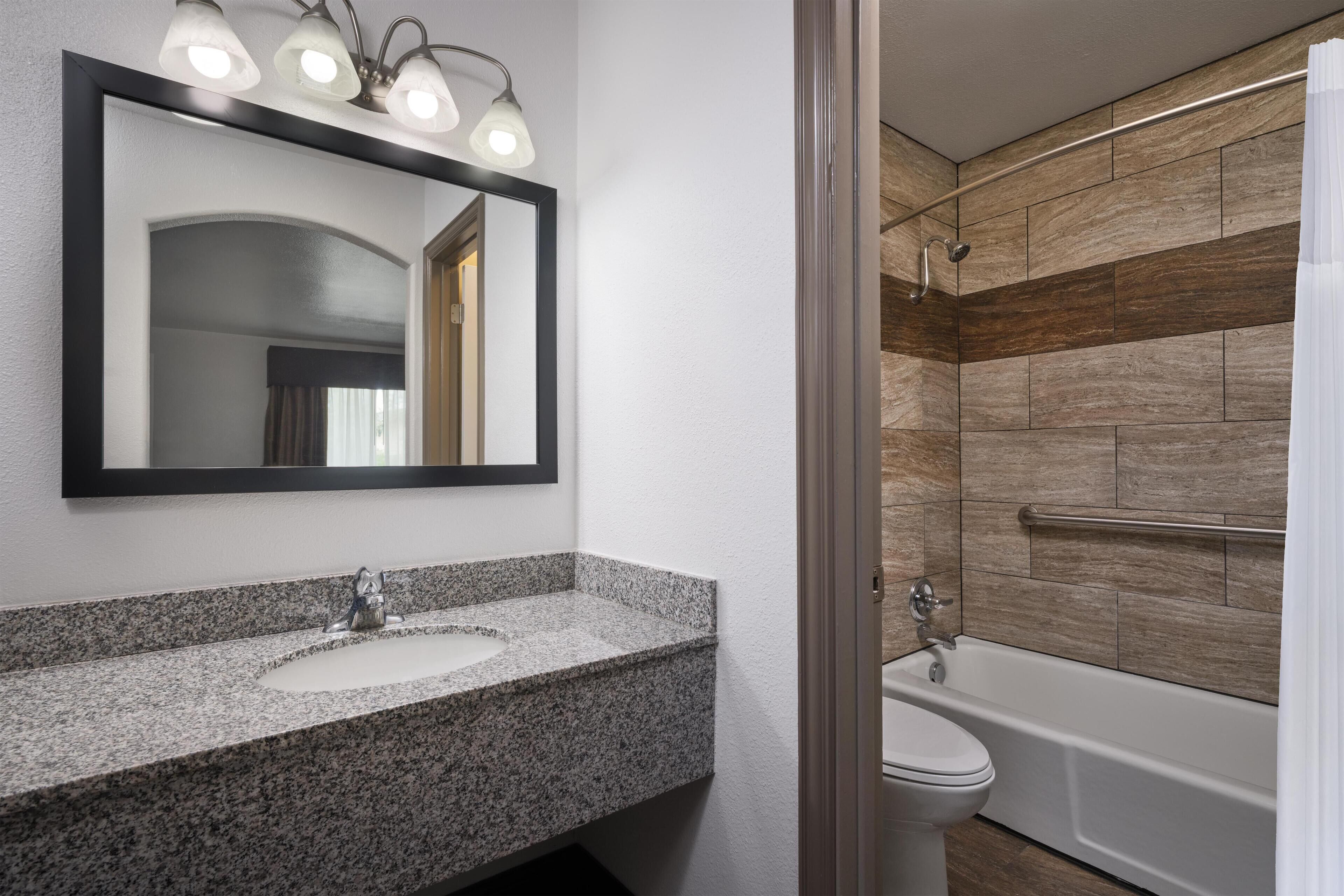 A bathroom with a granite countertop sink, a mirror with lights, a toilet, and a shower area with wooden tiles.