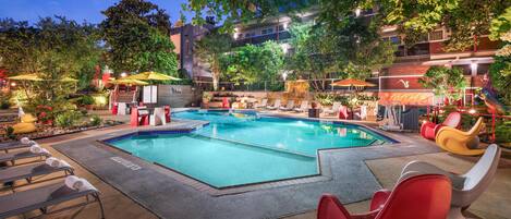 A swimming pool area with lounge chairs and umbrellas, surrounded by greenery and buildings.