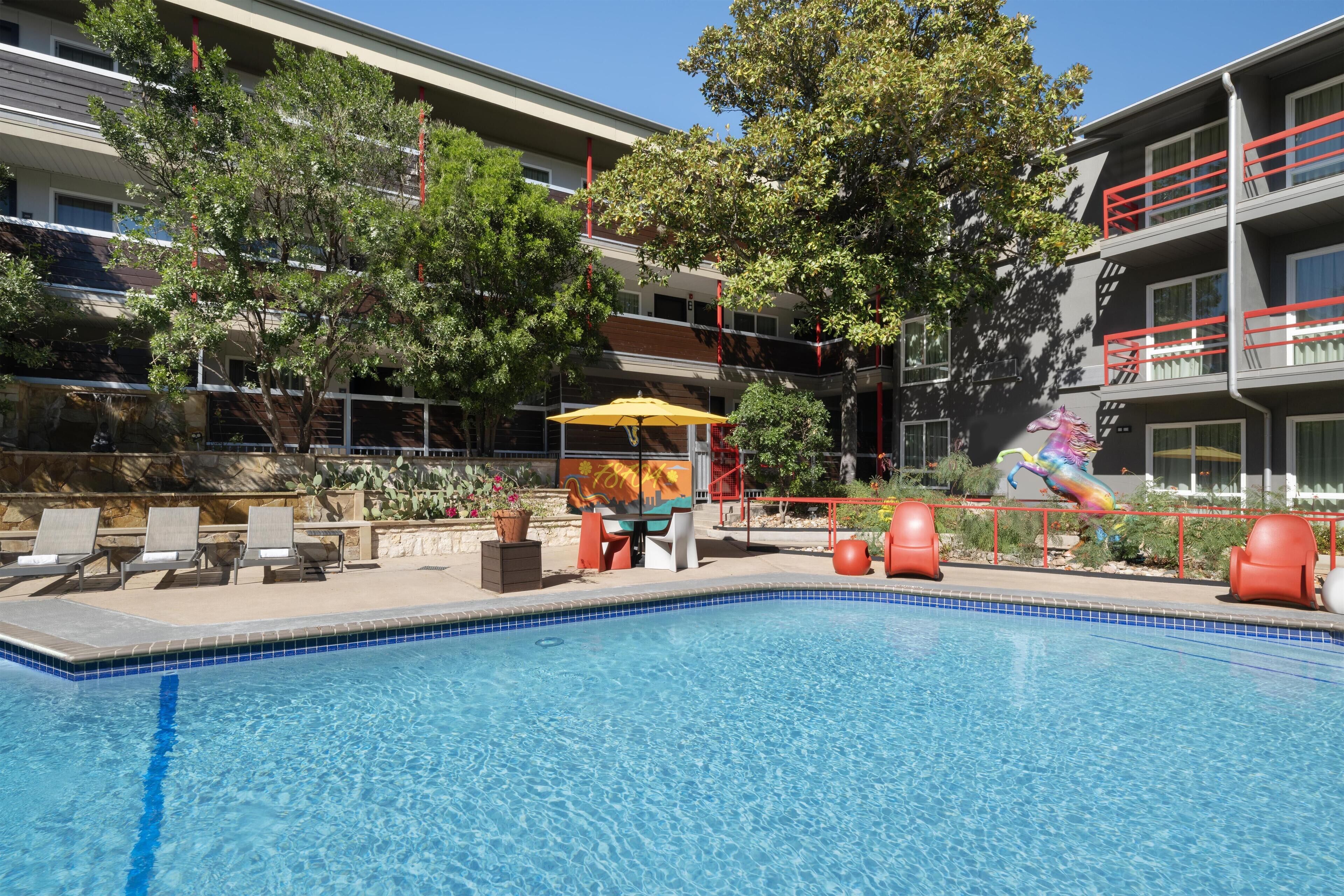 A swimming pool with lounge chairs and a sun umbrella, surrounded by trees and a building with balconies.