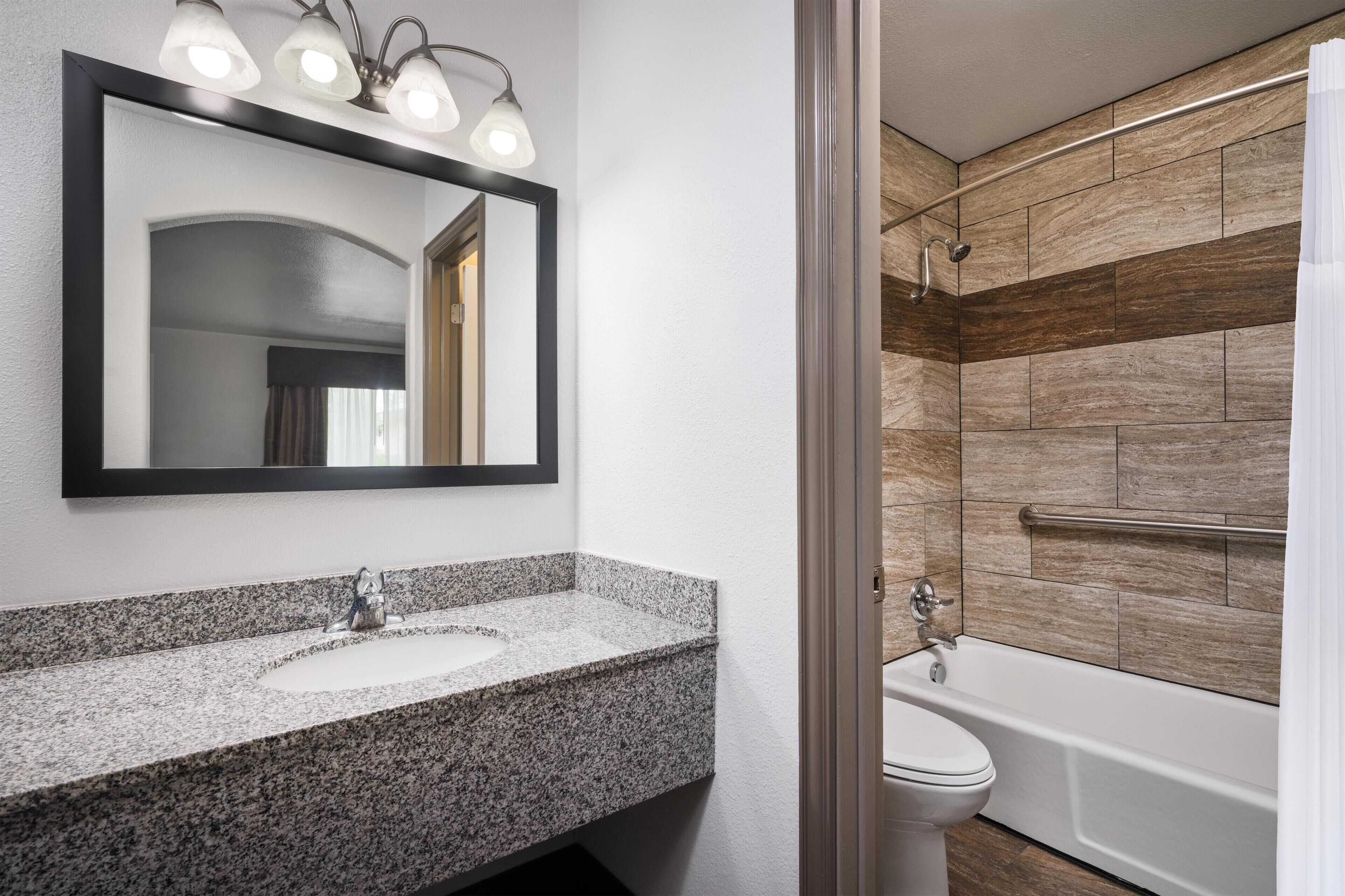A bathroom with a granite countertop sink, a mirror with lights, a white toilet, and a shower with wooden tiles.