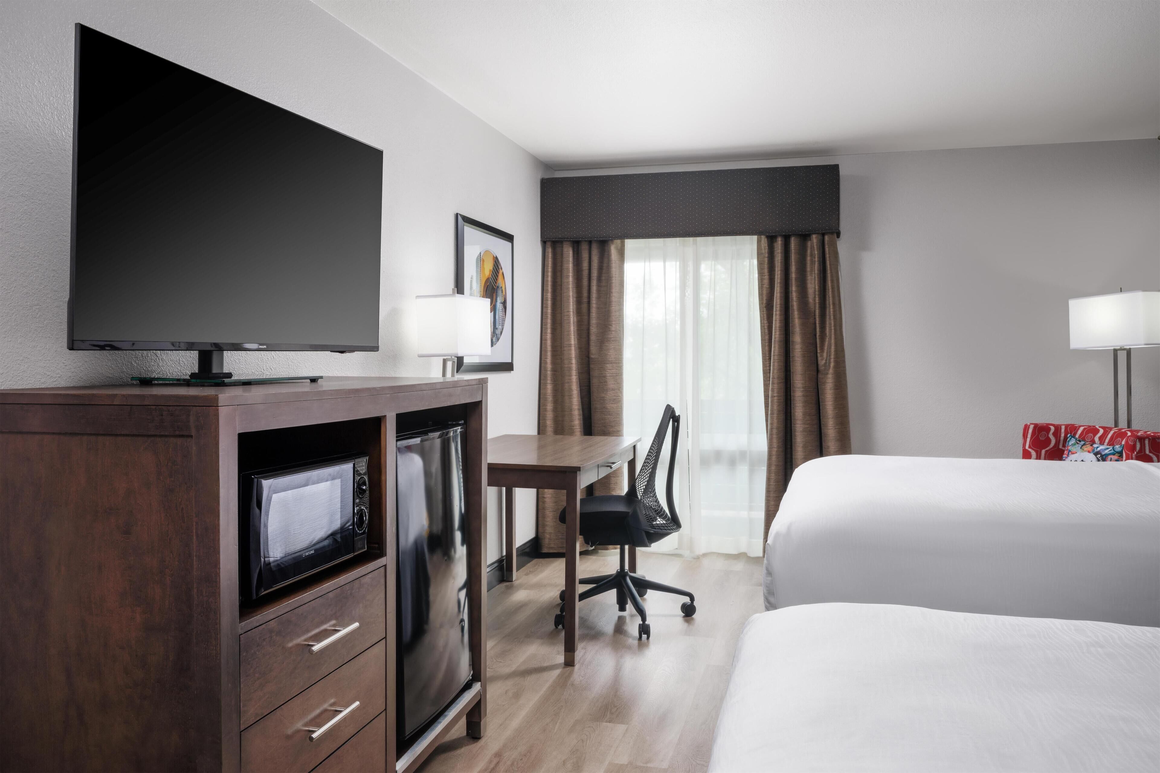 Habitación de hotel con televisión de pantalla plana, escritorio con silla, microondas, cama y ventana con cortinas.