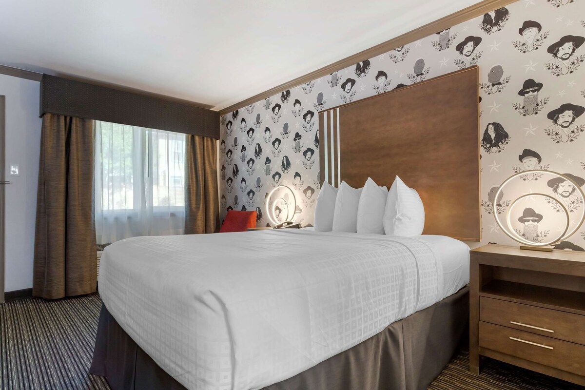 A hotel room with a large bed, a bedside table with a mirror, a wooden headboard, and patterned wallpaper.