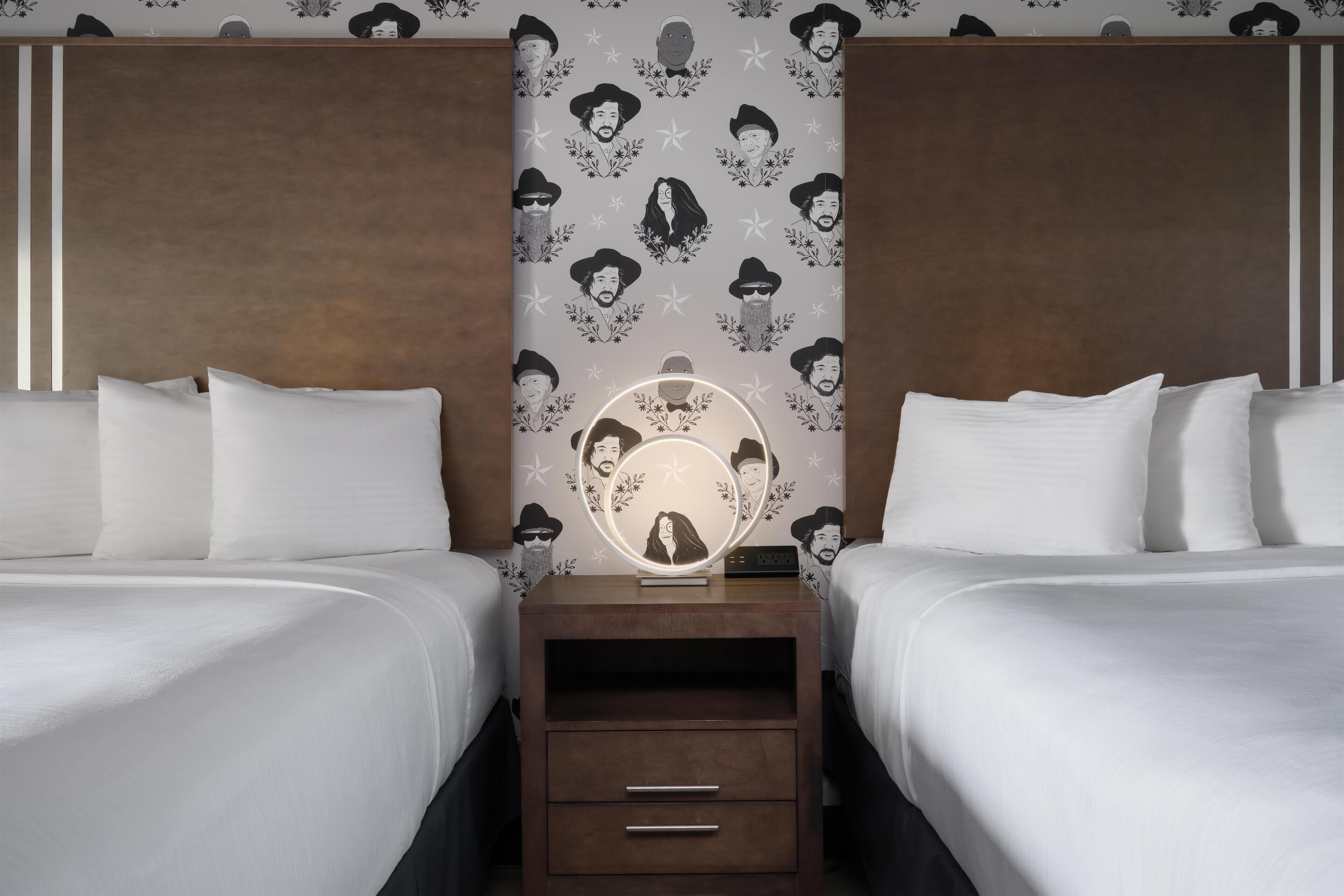 A hotel room with two beds, a nightstand with a mirror, and a wall with a pattern of faces.