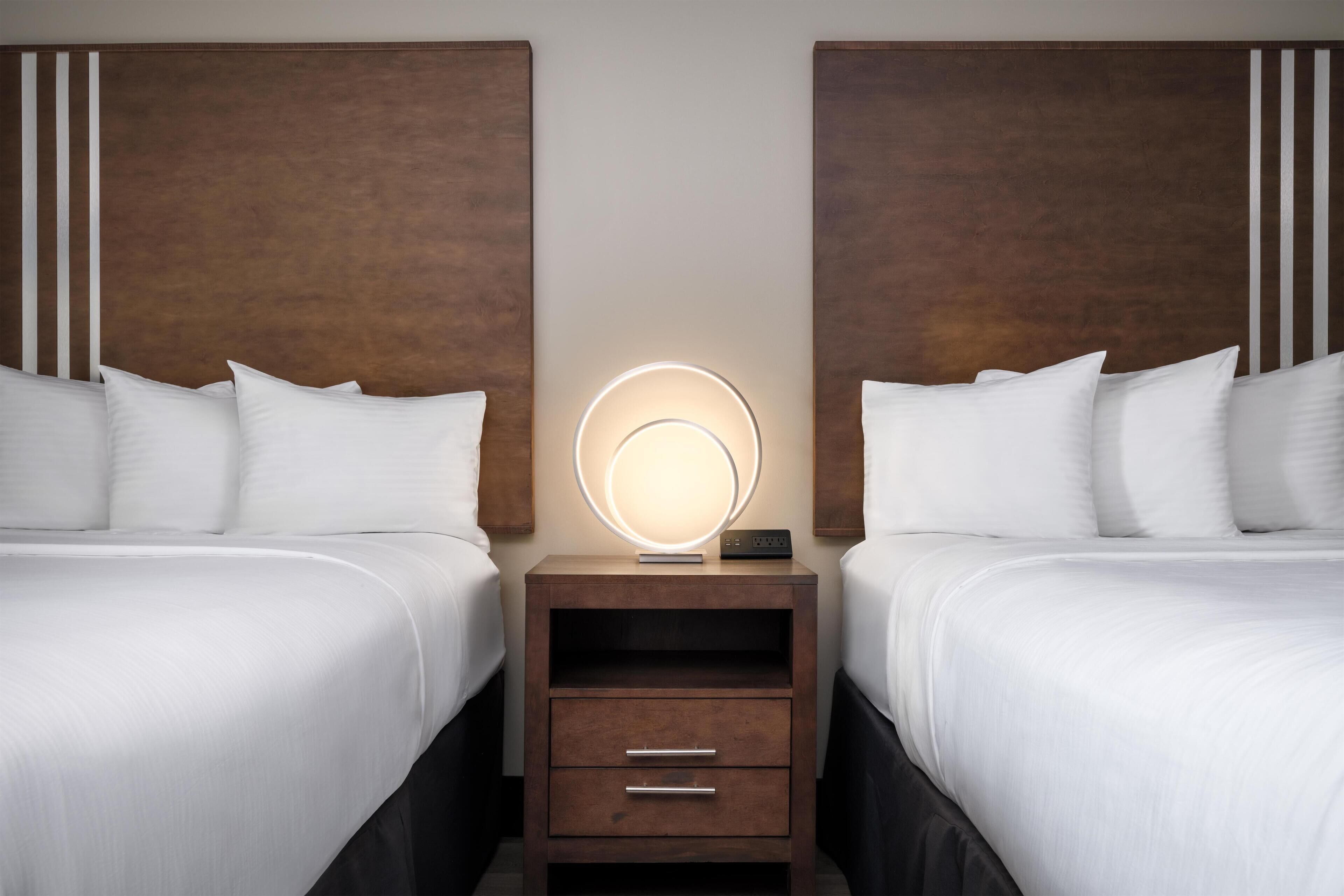 Habitación de hotel con dos camas, una mesita de noche con una lámpara circular y un cabecero de madera.