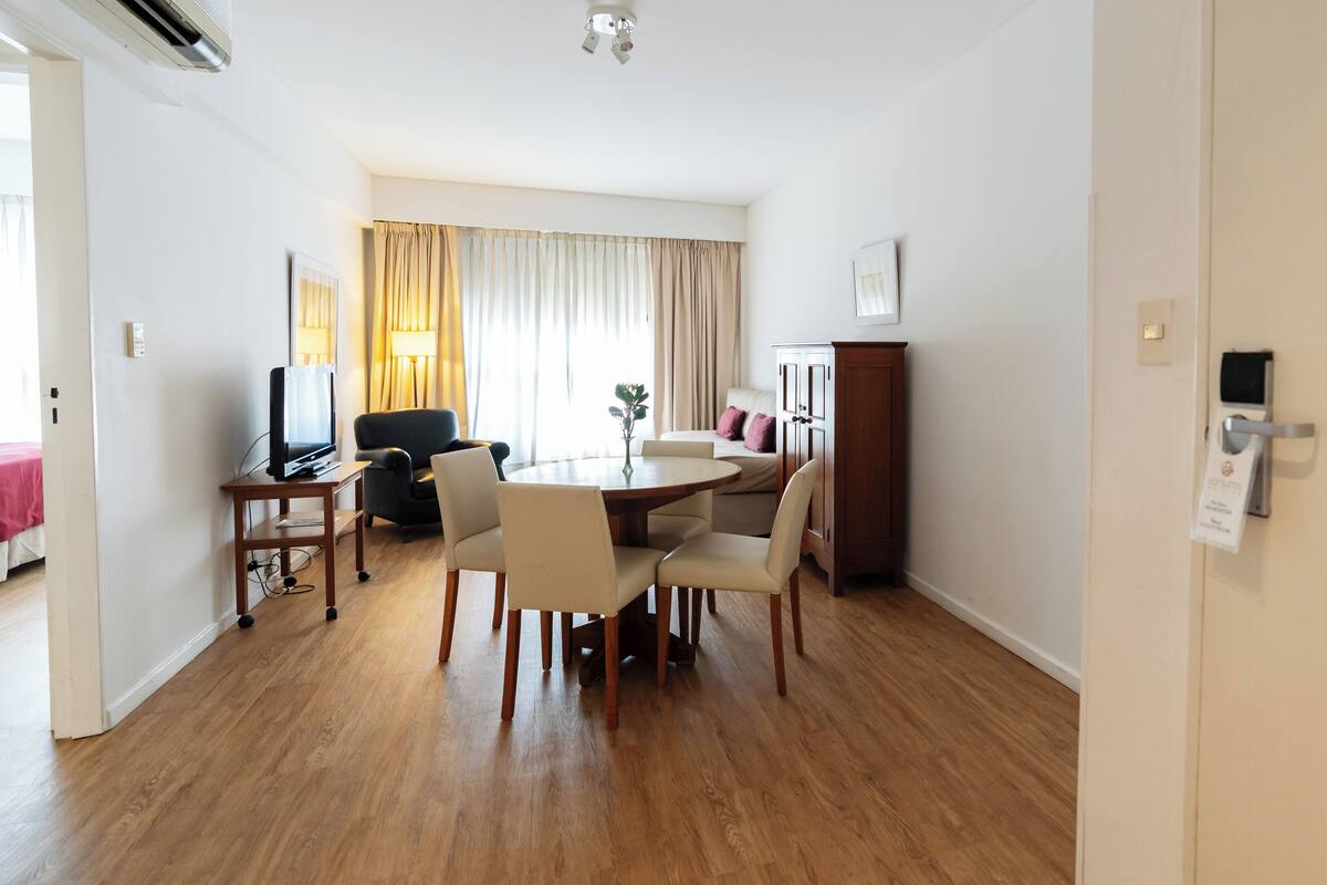 A hotel room with a dining area featuring a round table, four chairs, a TV on a stand, and a cabinet.