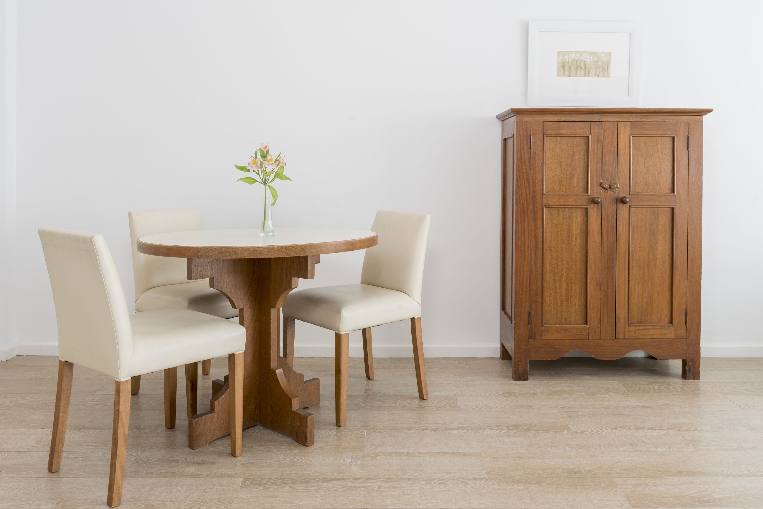 A dining table with two chairs, a vase with flowers, and a wooden cabinet against a white wall.