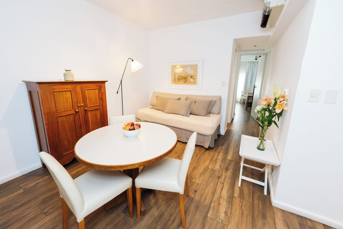 A dining area with a round table, two chairs, a wooden cabinet, a sofa, and a small side table with a vase.