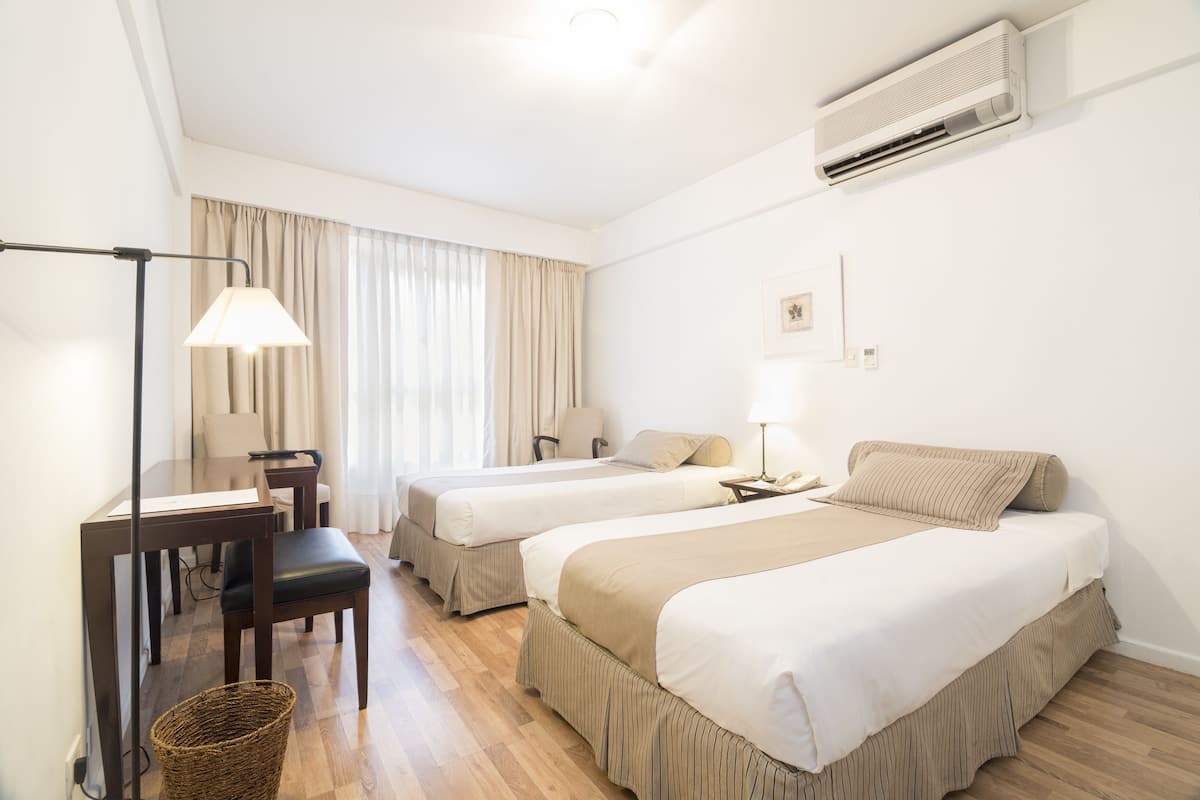 A hotel room with two beds, a desk, a chair, a lamp, and an air conditioning unit.