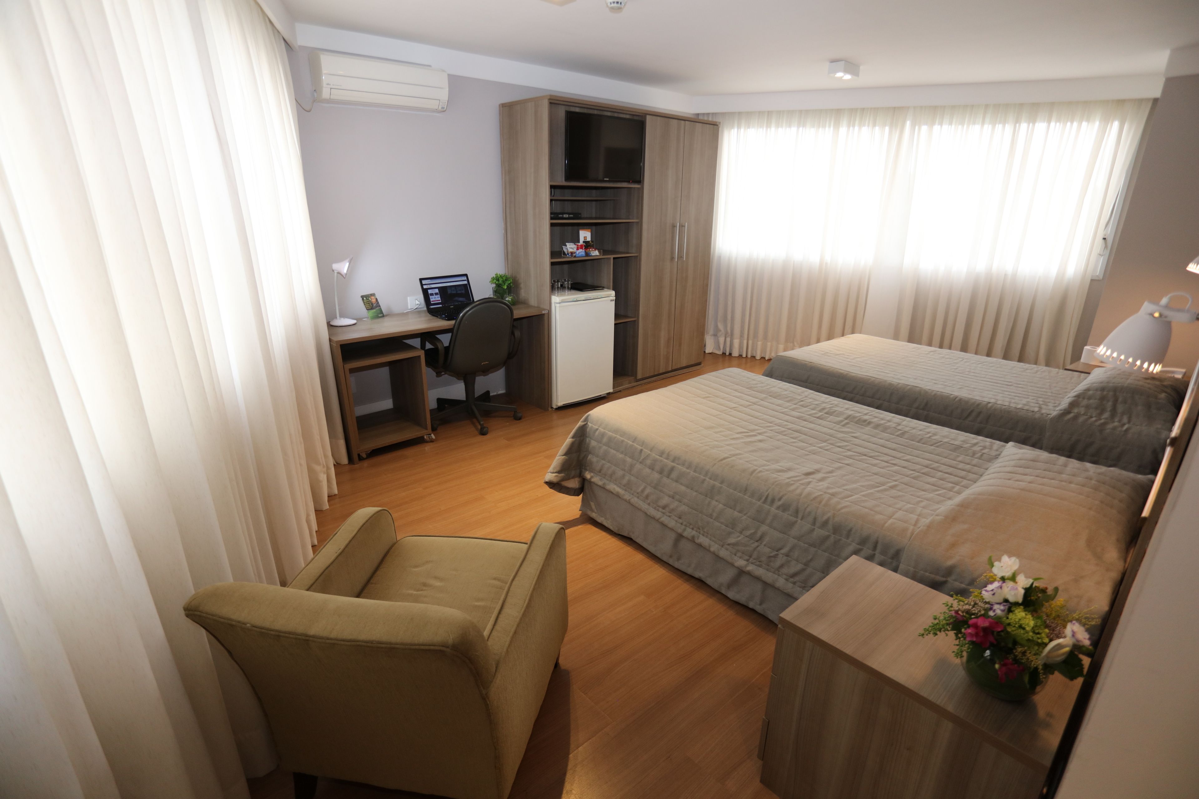 A hotel room with a bed, a desk, a chair, a wardrobe, and a small table with a vase of flowers.