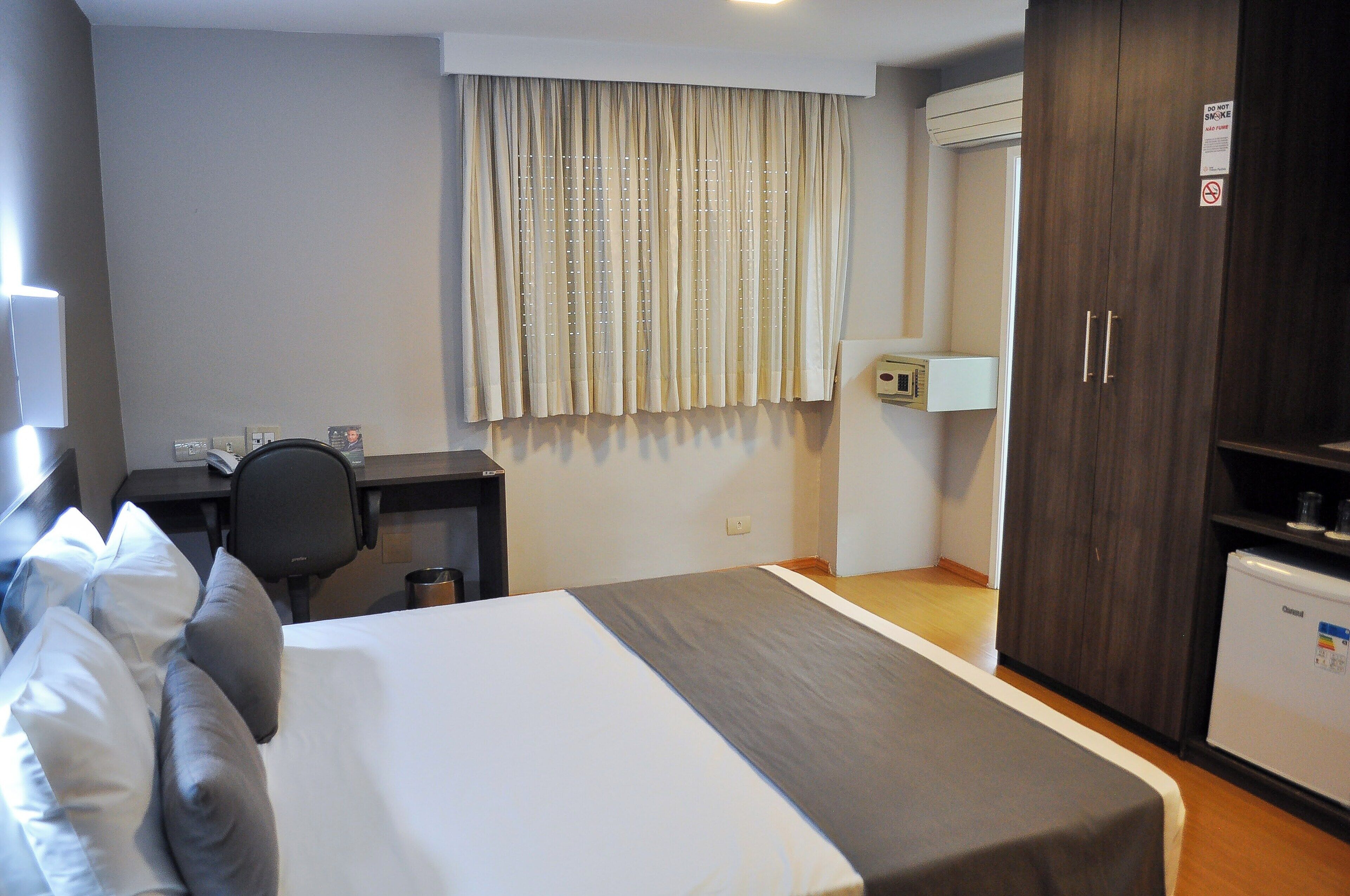 A hotel room with a bed, a desk, a chair, a wardrobe, and a mini-fridge.