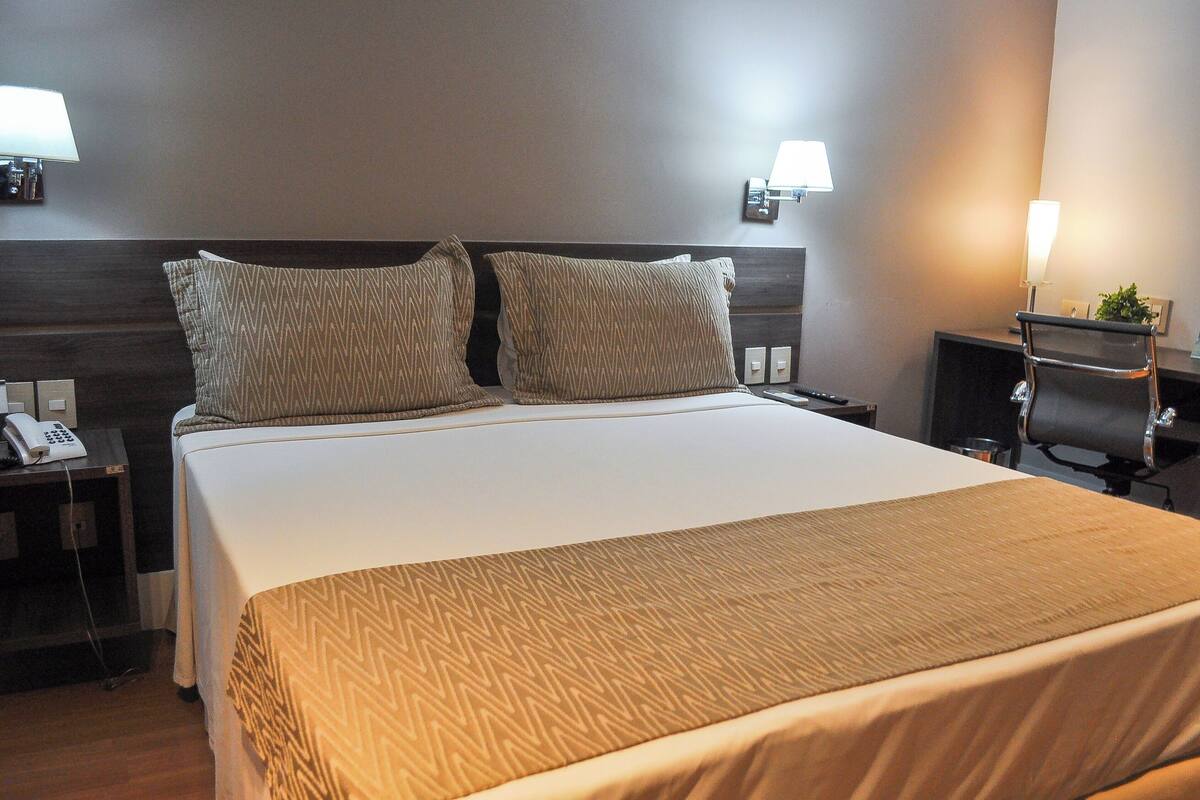 Une chambre d’hôtel avec un grand lit, deux oreillers, une table de chevet avec une lampe, un téléphone et une chaise.
