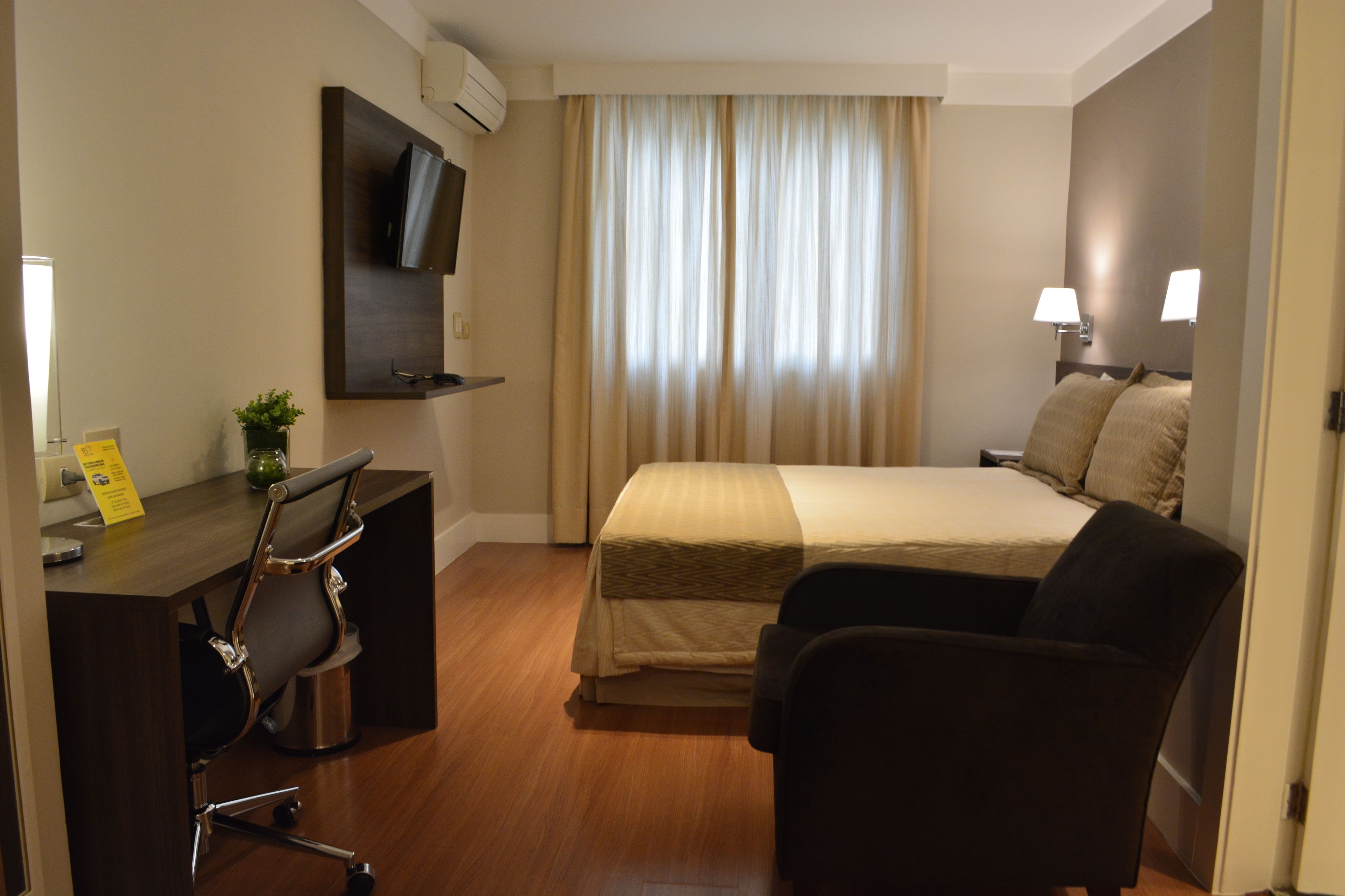 A hotel room with a bed, a desk, a chair, a TV, and a window with curtains.