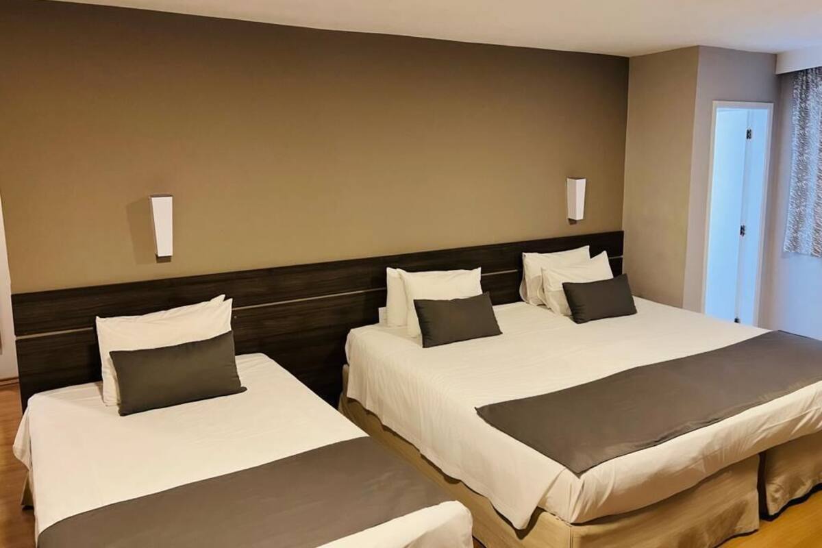 Une chambre d’hôtel avec trois lits, une tête de lit en bois et un mur sobre.
