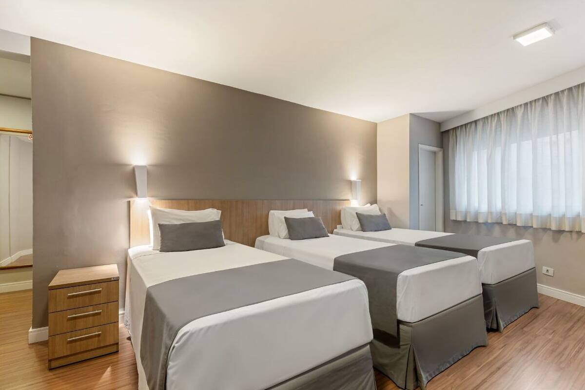 Une chambre d’hôtel avec trois lits simples, une table de chevet en bois et une grande fenêtre avec des rideaux légers.