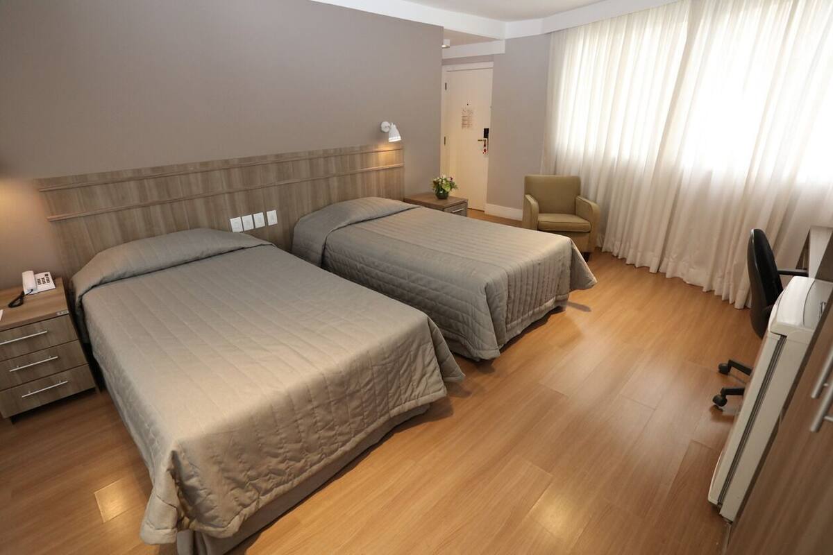 Une chambre d’hôtel avec deux lits, un bureau, une chaise et une grande fenêtre avec des rideaux.