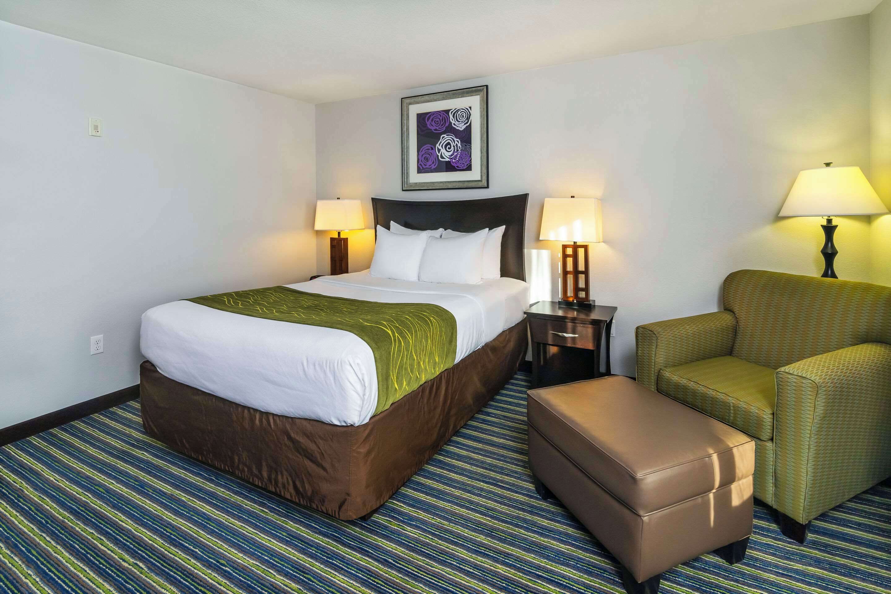 Une chambre d’hôtel avec un grand lit, deux lampes de chevet, un fauteuil vert, un pouf marron et une œuvre d’art encadrée au mur.