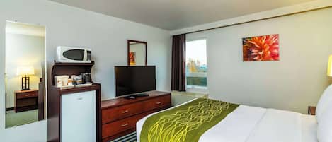 Habitación de hotel con cama, televisor, refrigerador pequeño, escritorio con microondas y ventana con cortinas.