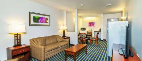 A hotel room with a sofa, a coffee table, a TV, a refrigerator, a dining area with chairs and a table, and a kitchen area with a microwave.