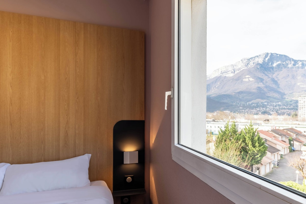 Ein Hotelzimmer mit einem hölzernen Kopfteil, einem Nachttisch mit Lampe und einem Fenster mit Blick auf Berge und Gebäude.