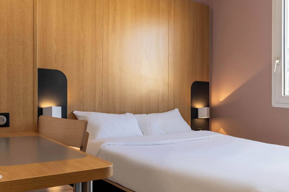 Ein Hotelzimmer mit massivem Holzkopfteil, einem Bett mit weißen Leinentüchern, einem Nachttisch und zwei Wandleuchten.