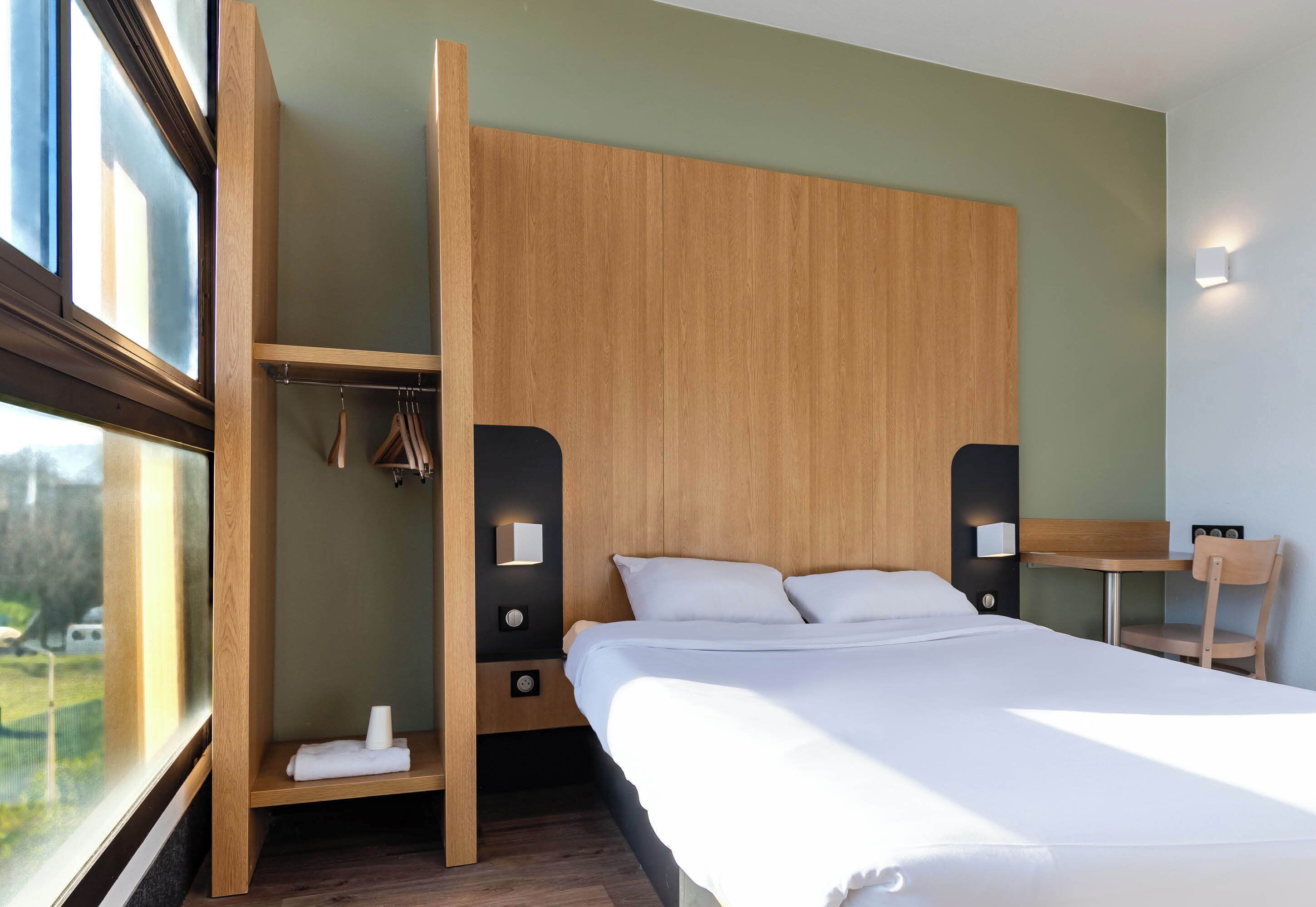 A modern hotel room with a large bed, a wooden wardrobe, a small table, and a chair.