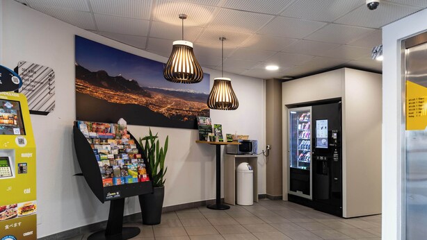 A reception area with a large poster of a cityscape, vending machines, a trash bin, and a microwave on a small table. - B&B HOTEL Grenoble Centre Verlaine (Grenoble)
