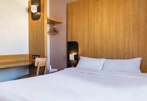 A hotel room with a wooden headboard, a bed with white linens, a wooden desk, a chair, and a wall-mounted light fixture.