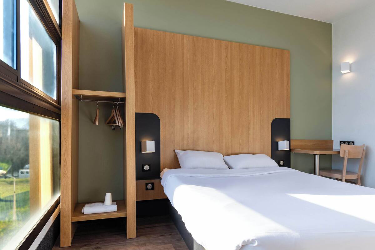Ein modernes Hotelzimmer mit einem großen Bett, einem Holz-Kopfteil, einer kleinen Sitzecke und Blick durch ein Fenster.