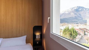 A hotel room with a wooden headboard, a bedside table with a lamp, and a window offering a view of a mountain and buildings. - B&B HOTEL Grenoble Centre Verlaine (Grenoble)