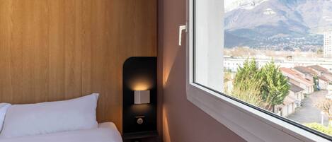 Habitación de hotel con cabecera de madera, mesita de noche con lámpara y ventana con vistas a una montaña y edificios.