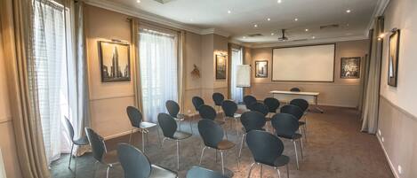 Un salón de conferencias con filas de sillas, una pantalla de proyector y cuadros en las paredes.