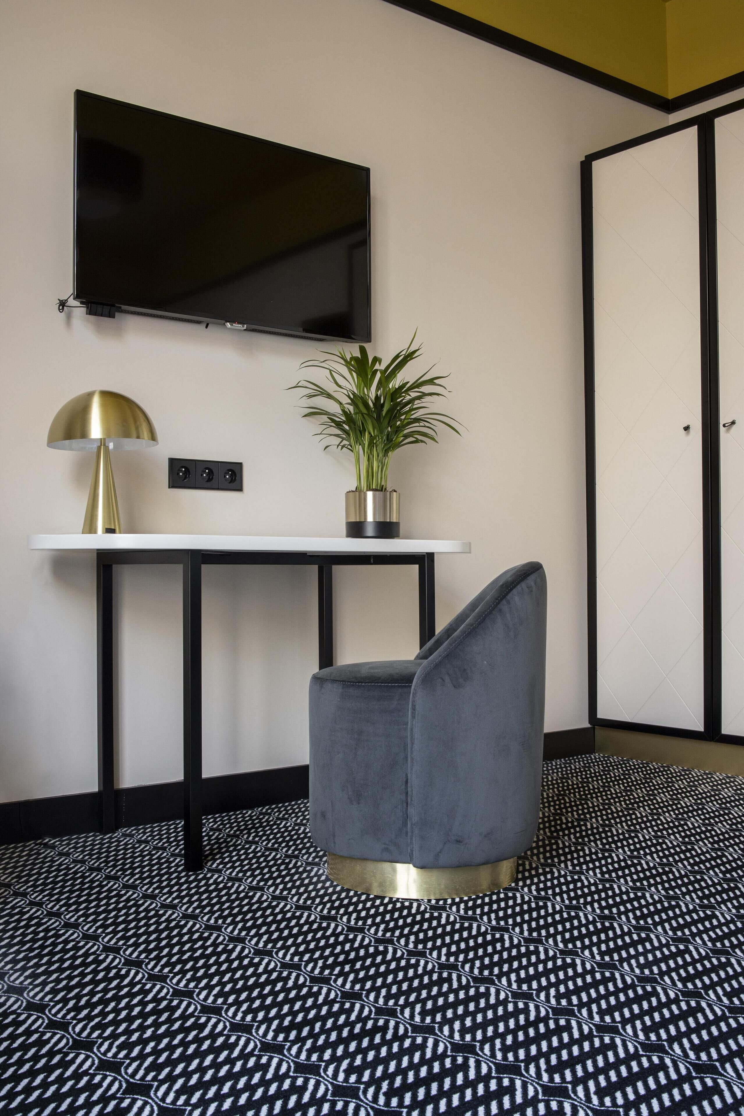 A modern room with a flat-screen TV mounted on the wall, a small table with a potted plant, a grey upholstered chair, and a patterned floor.