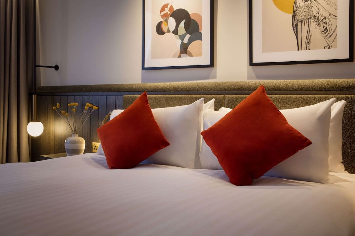 Hotellihuone, jossa on sänky, kaksi punaista tyynyä, maljakko, jossa on keltaisia kukkia, ja kaksi kehystettyä taideteosta seinällä.
